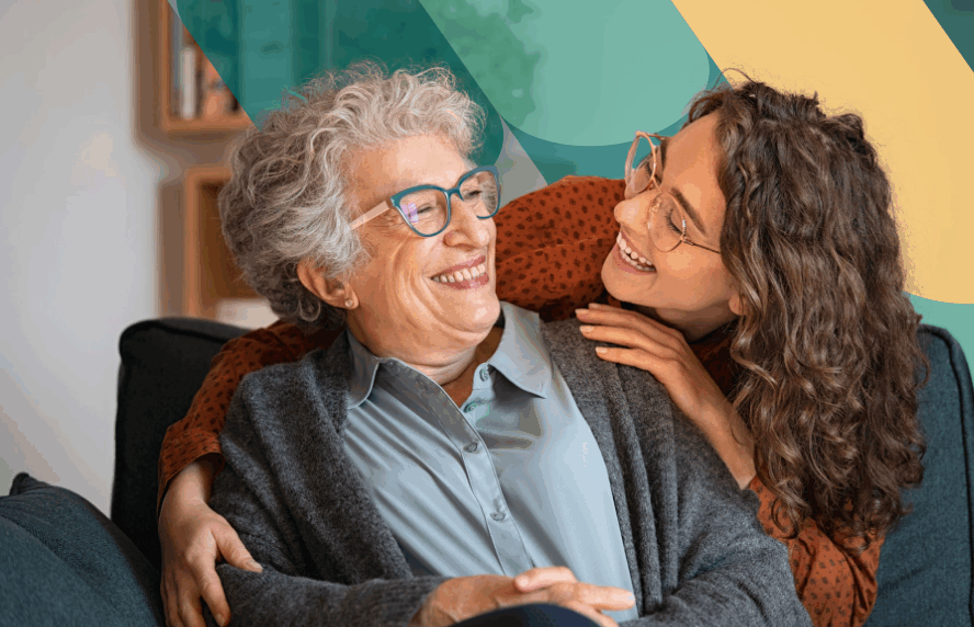 Young white woman with brown wavy hair and glasses leaning over couch hugging older white woman with short curly hair and glasses who is sitting on couch