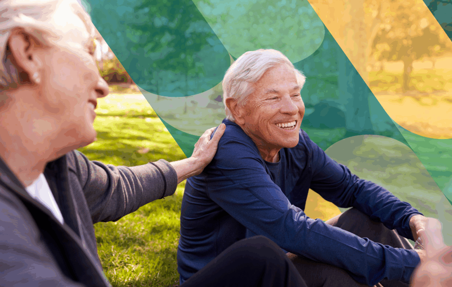 Senior woman reaching out and patting the back of a senior man, both smiling and sitting in the grass