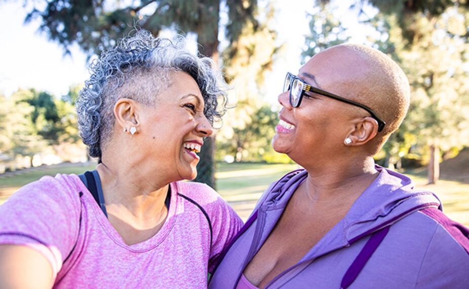 Two women smile and embrace each other in a grassy park. One wears a pink shirt and has gray, curly hair, and the other has a purple button-down and black glasses, and is bald.