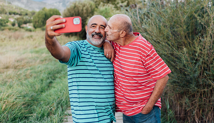 Two men embrace and take a selfie together in a green, park-like setting. One is wearing a green striped shirt and has gray facial hair. The other is wearing a red stripe shirt and is giving the first a kiss on the cheek.