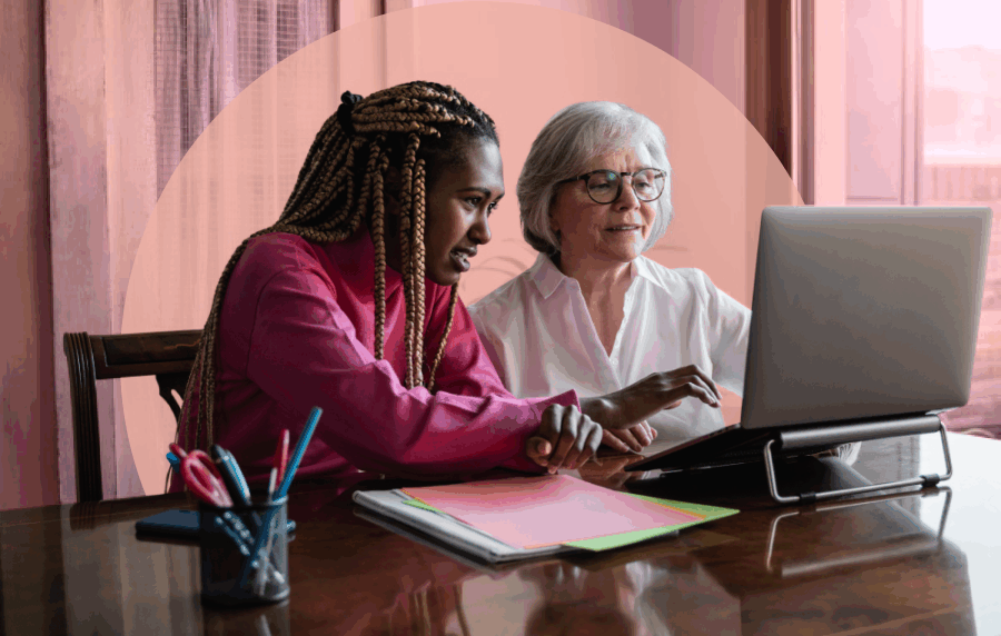 Two women sitting together at a table and looking at a laptop screen - a younger black woman with long hair and an older white woman with short white hair and glasses.