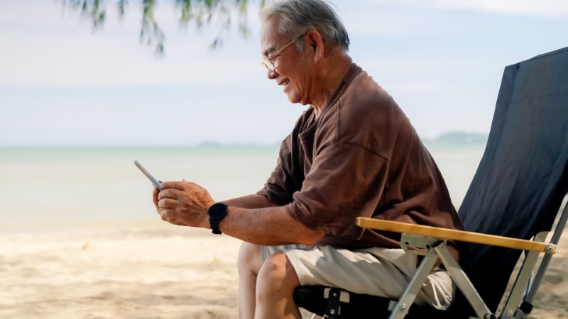 Senior man at the beach sitting a lounge chair, looking at his cell phone and smiling