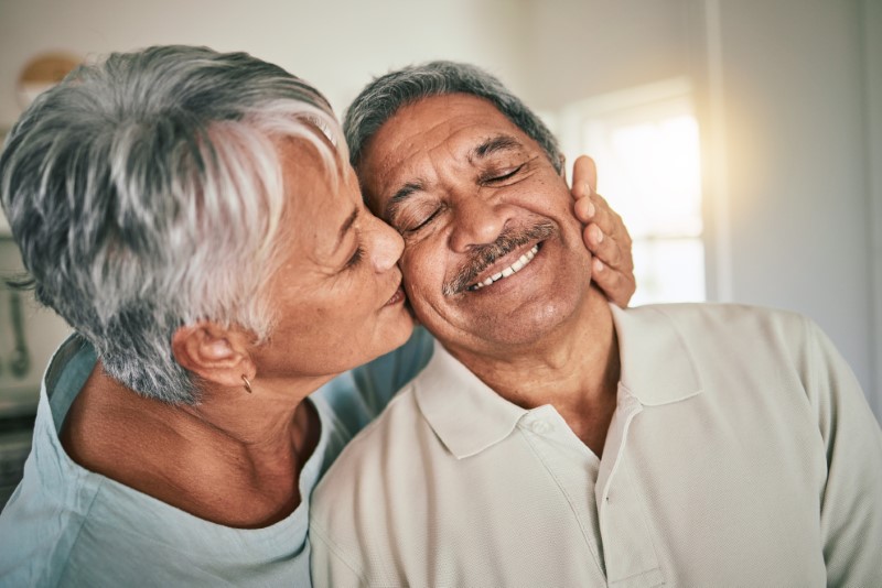 Happy older couple smiling smiling while woman gives her husband a kiss on the cheek.