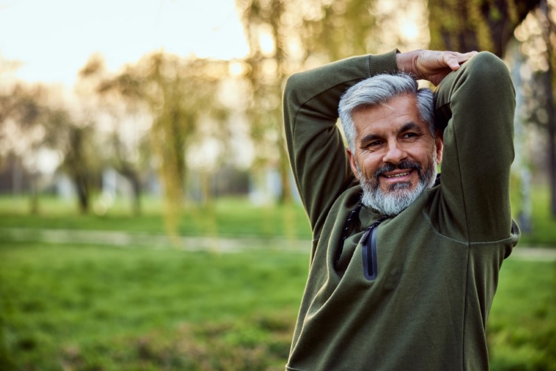 A cheerful senior bearded man enjoying himself in nature, sitting on a bench and looking around the park.