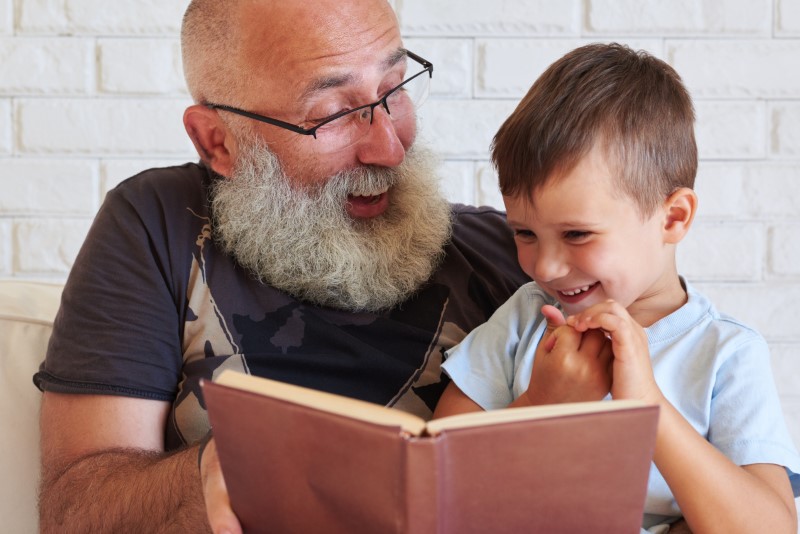 Portrait of grandfather and his small grandson reading a book together on couch at home