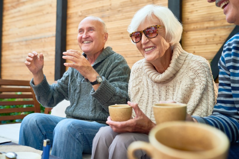Cheerful senior friends gathered together at cozy small patio and remembering funny stories from their past, they wearing knitted sweaters and warming themselves with tea