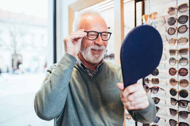 Senior man choosing eyeglasses frame in optical store, smiling while looking in handheld mirror