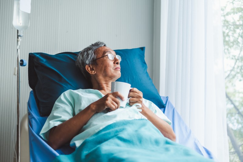 Older Asian male patient in bed in hospital with a coffee cup in hand, looking out window