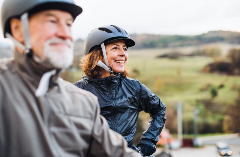 An active senior couple with helmets and bikes standing outdoors on a road in nature.