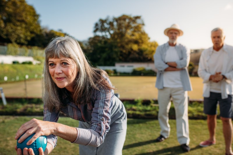 Close up of a senior woman playing boules with her male friends standing in the background. She is bending forward to throw a boules in a park.