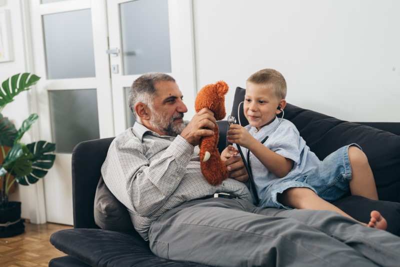 boy and his grandfather sitting on sofa and playing with a stuffed animal and stethoscope at home