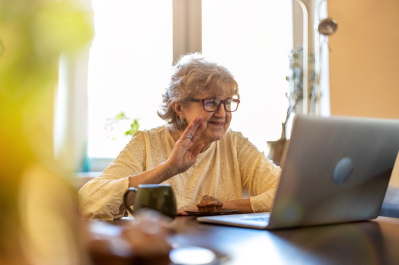 Elderly woman on a video call sitting at kitchen table, waving at screen.