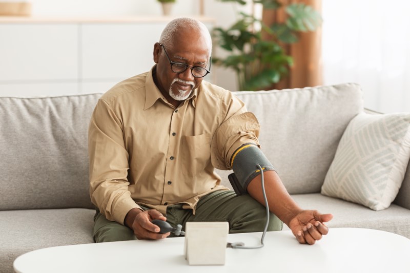 African American Man Measuring Blood Pressure Sitting On Couch At Home.