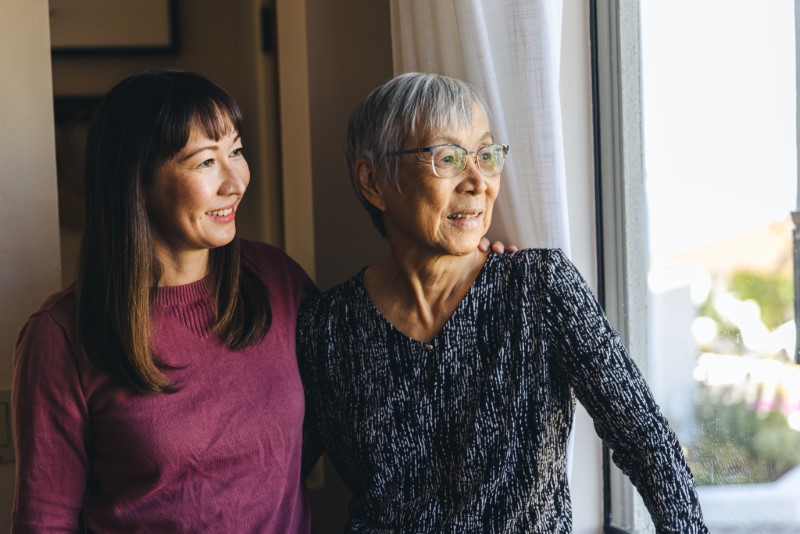 Smiling mature woman with arm around mother looking through window at home