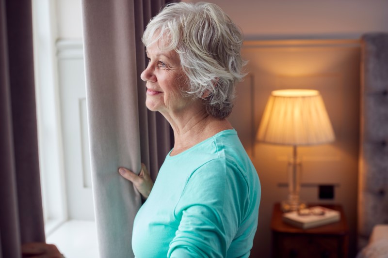 Smiling Senior Woman At Home Opening Bedroom Curtains And Looking Out Of Window