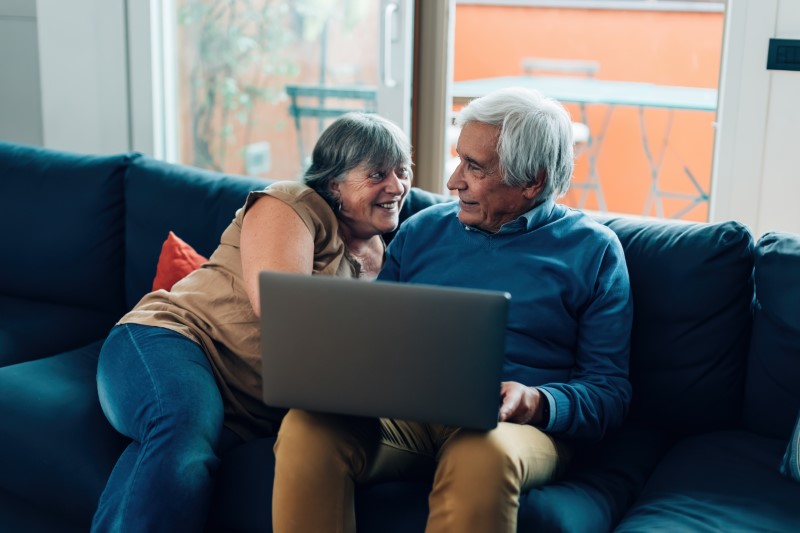 elderly couple sitting on sofa using laptop and smiling at one another