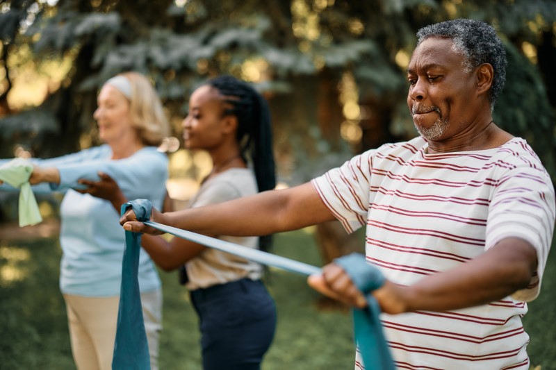 African American senior man using resistance band during exercise class in a green space. A young woman helps a senior woman stretch in the background.