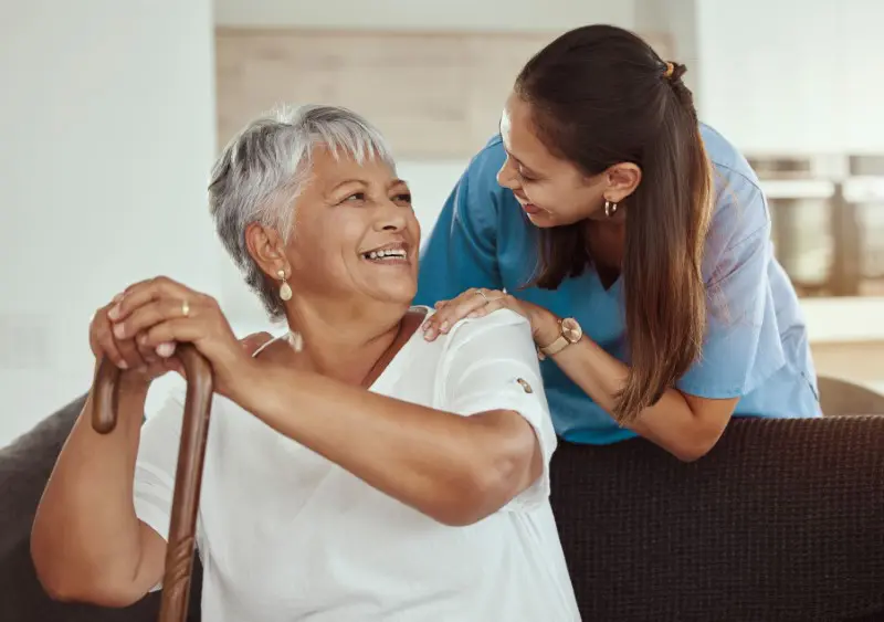 Senior woman with caregiver smiling while sitting on a living room sofa in a nursing home.