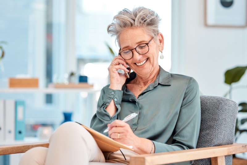 An older business woman talking on her phone while smiling and writing an appointment in her notebook.