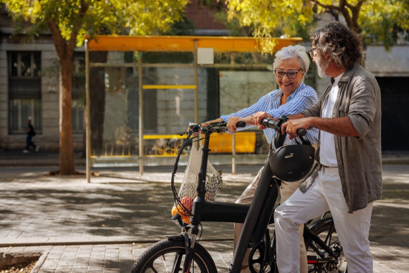 A cheerful mature couple is out in the city streets talking to each other and using an electric scooter and bike.