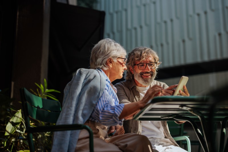 A stylish senior couple is outside in a garden of a beautiful cafe on a sunny day using their smartphone.