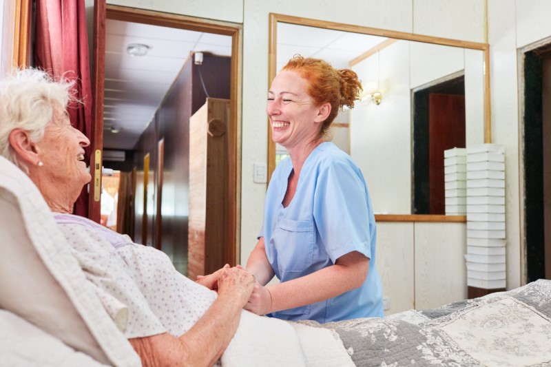 Laughing geriatric nurse sits at the bedside of a sick elderly woman and comforts her