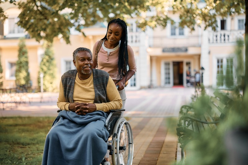 Young black woman pushes her senior father in wheelchair while visiting him at his nursing home.