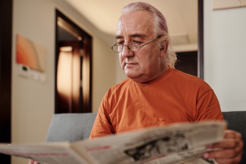 Aged man in glasses sitting in couch at home and reading newspaper