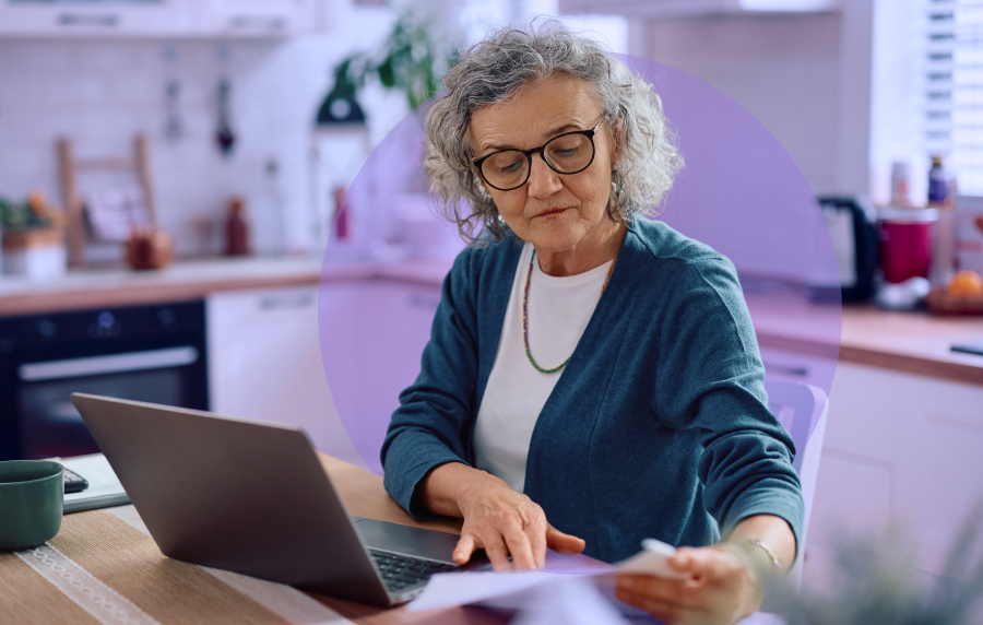 Senior woman with curly salt-and-pepper hair and glasses sitting at a kitchen table with a laptop in front of her, looking at documents