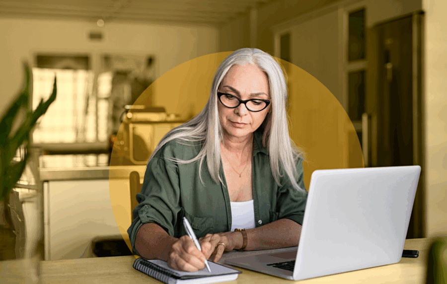 Seniors woman with long white hair and glasses sitting at dining room table and taking notes, with a laptop open in front of her