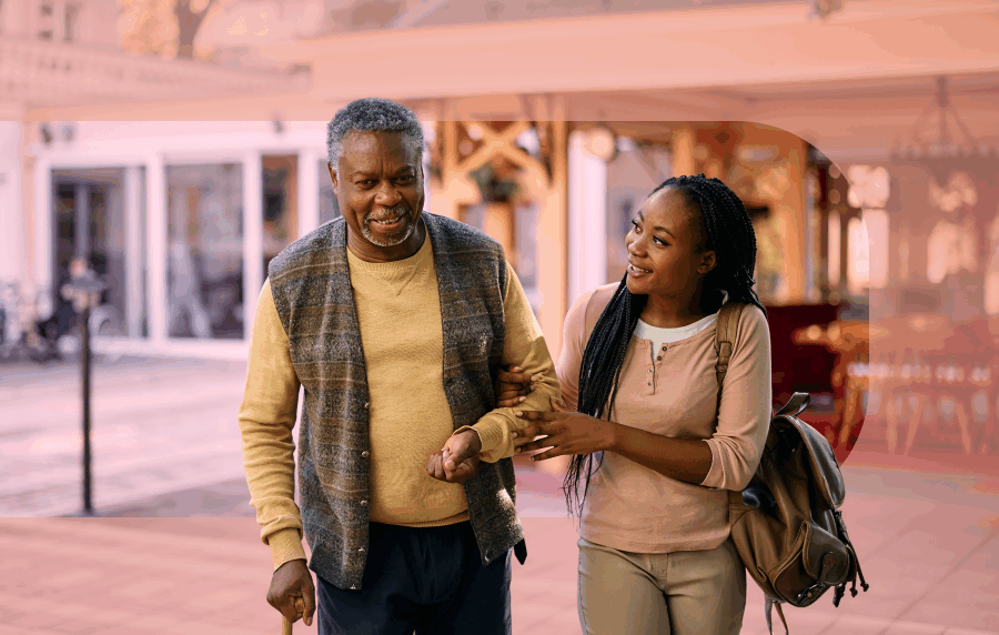 Older Black man walking with a cane being helped by a younger Black woman who is smiling at him