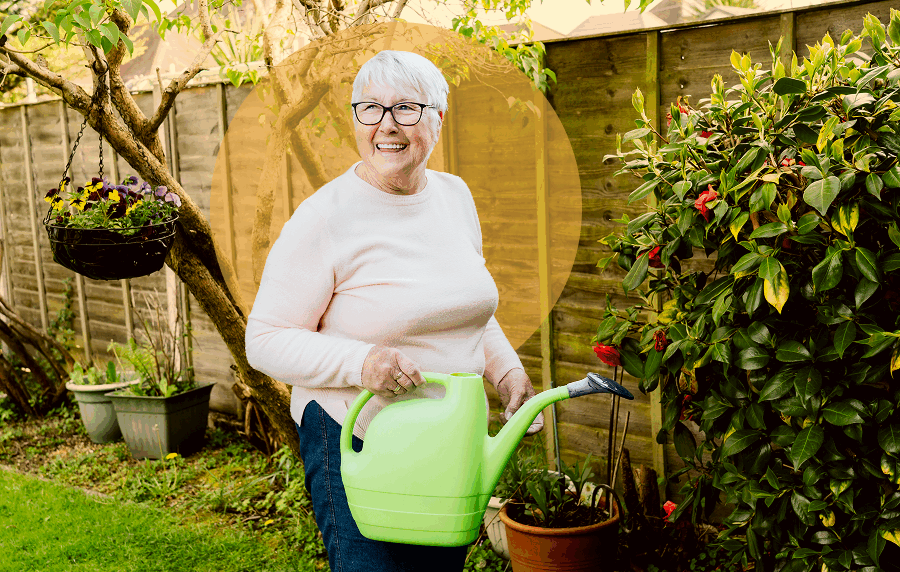 Older white woman with short hair in a bakyard, smiling and holding a watering can