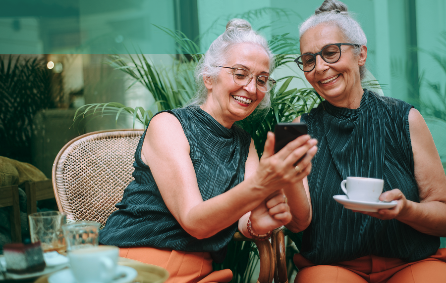Two seniors women sitting at an outdoor cafe looking at the screen of a phone.. Both women are white with white hair in buns and wearing glasses, and one is holding a cup and saucer.