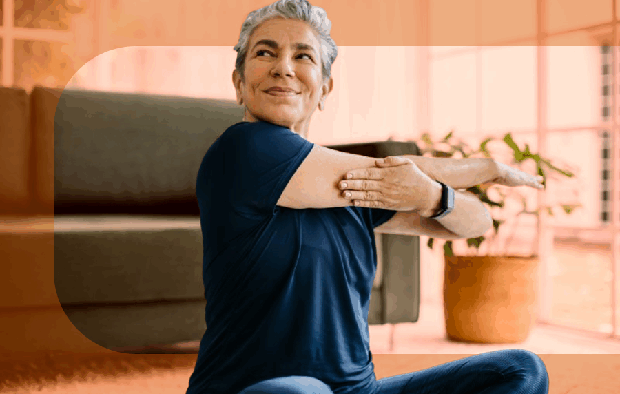 Middle-aged woman sitting on the floor in a living room, stretching one arm across her chest and smiling