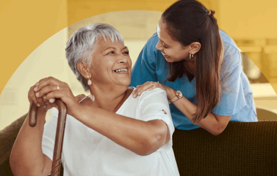 Two Hispanic women, a senior woman sitting on a couch and holding a cane, smiling up at a younger woman in scrubs