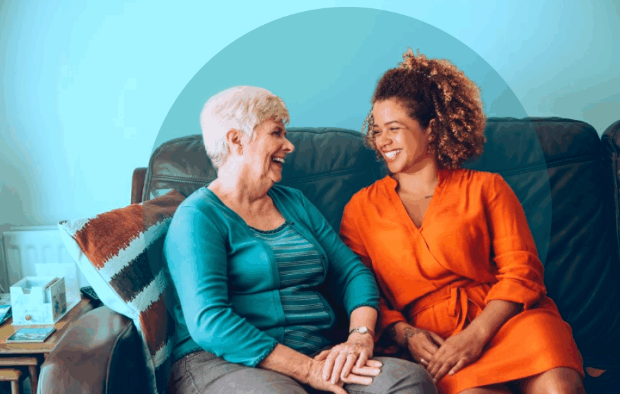 Two women sitting on a couch and smiling at one another - a younger black woman and senior white woman