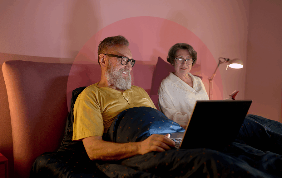 Senior white couple in bed, both wearing glasses and looking at a laptop sitting on the man's lap