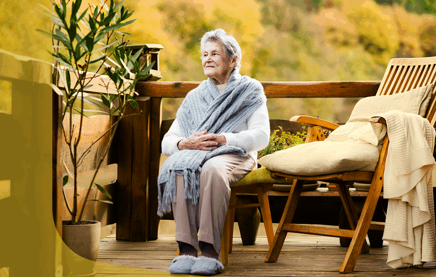 Elderly woman wrapped in shawl and wearing slippers sitting on a porch surrounded by greenery. She is gazing off into the distance.
