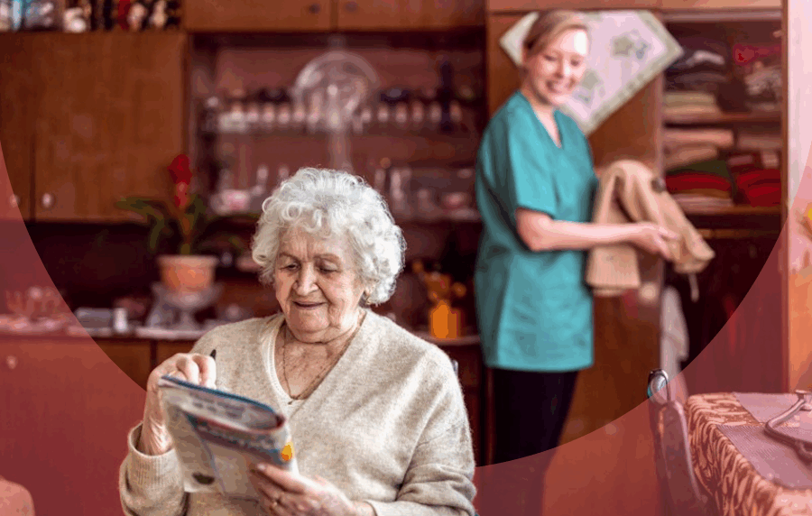 Elderly woman sitting in foreground looking at a magazine, with young woman in scrubs int he background looking towards her and smiling