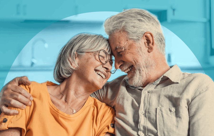 Close-up of white senior couple sitting together on couch and smiling, their foreheads pressed together and eyes closed