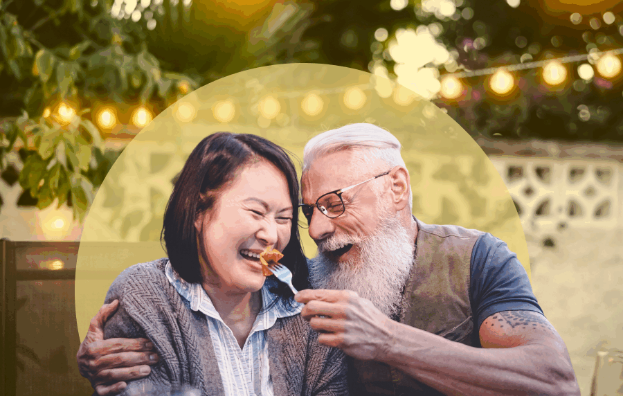 Senior couple - a white man and East Asian woman, dining outside. The man has white hair and a long white beard, and is feeling a bite of food to the woman, who is smiling.