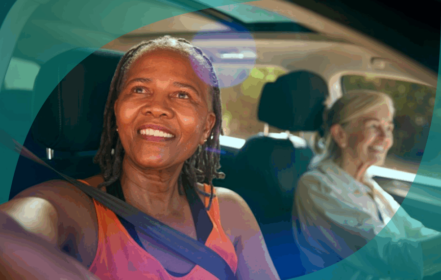 Two senior women in a car, a white woman in the driver's seat in the background and a smiling Black woman looking out the window in the foreground