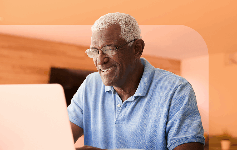 Black senior man in a blue polo shirt sitting at table smiling while looking at a laptop