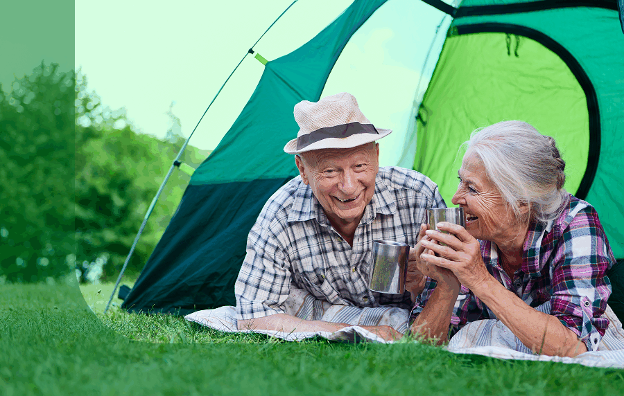 White senior couple camping, laying in the grass in front of a tent and smiling