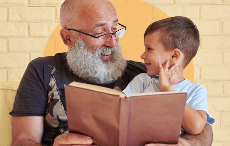 Senior man with bald head, white beard and glasses, reading to young boy sitting on his lap and smiling