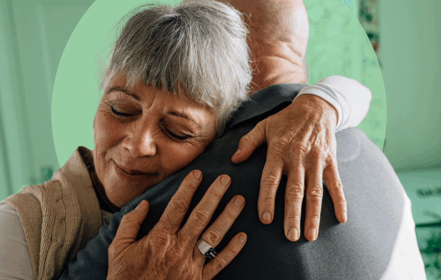 Close-up shot of two seniors hugging, showing the back of a man the front of a woman, whose face is in a soft smile with closed eyes