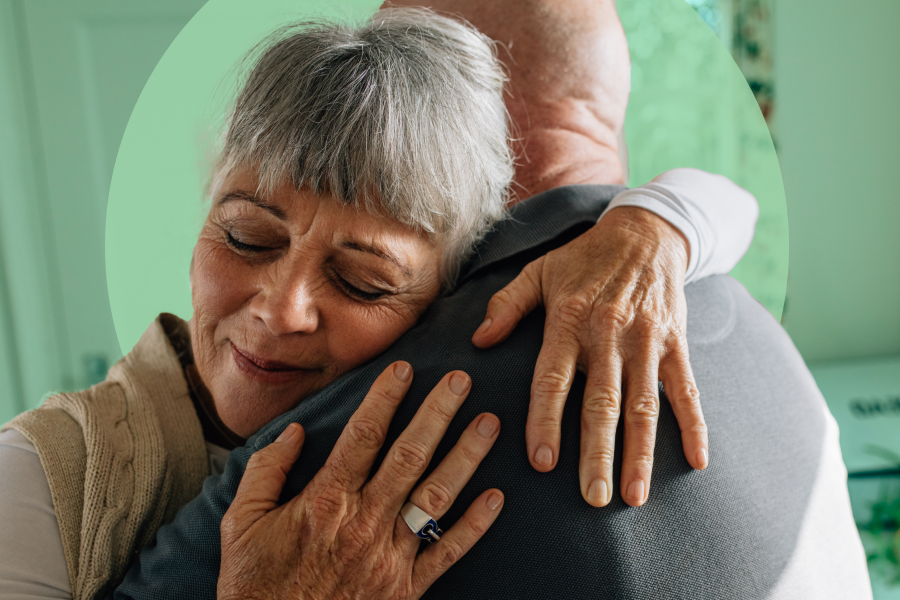 Close-up shot of two seniors hugging, showing the back of a man the front of a woman, whose face is in a soft smile with closed eyes