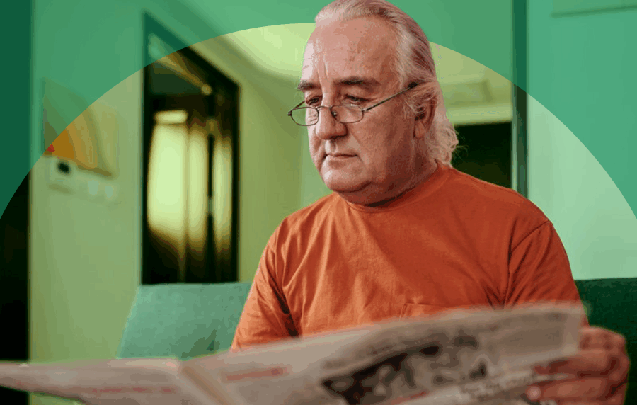 Senior man with white hair and glasses sitting on a couch reading a newspaper