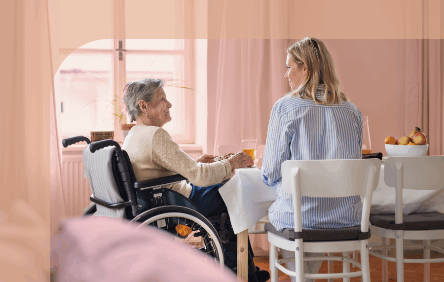 Senior woman in wheelchair sitting at dining table with younger woman with blode hair
