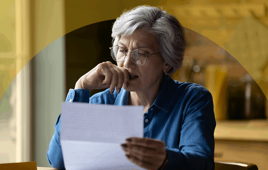 Senior woman with short white and grey hair and glasses sitting at a table reading a document, looking slightly uncertain
